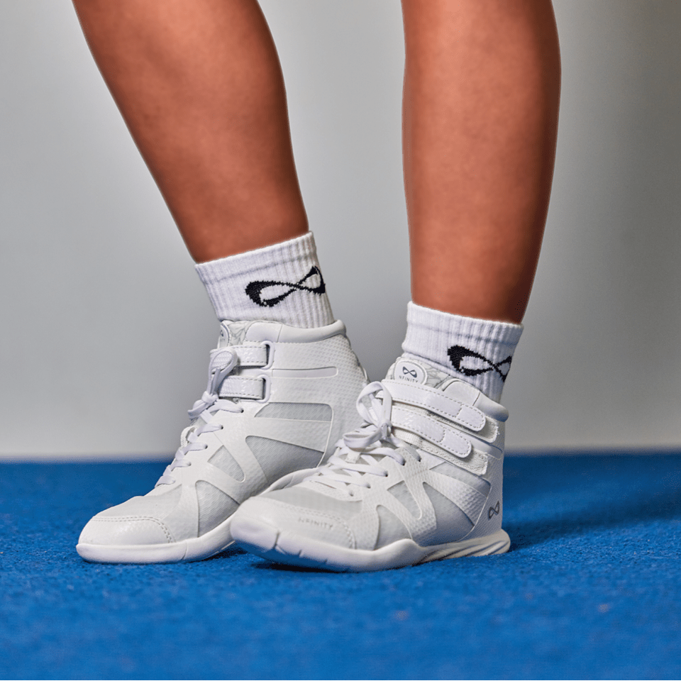 Cheerleader standing on a blue mat in white Nfinity Beast mid-top cheer shoes